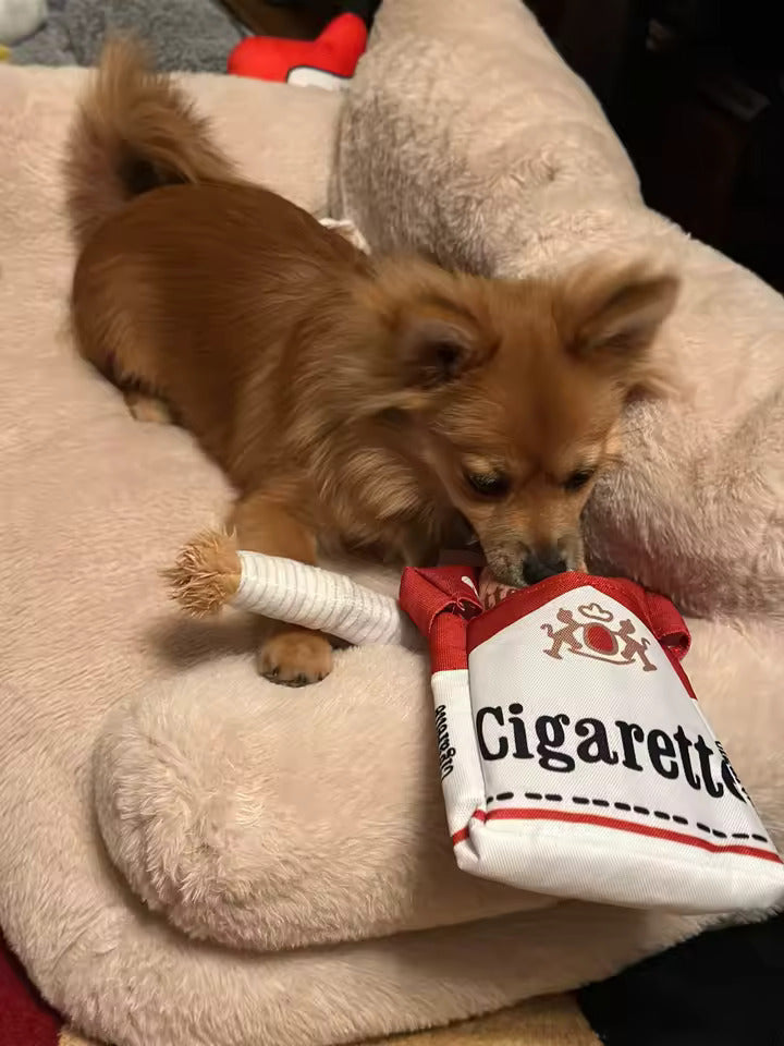 Small dog playing with a cigarette toy beside its plush cigarette box